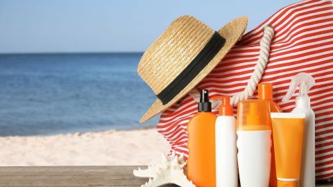 Sonnencremes und ein Strohhut sind auf einem Holztisch vor einem Strandarrangement platziert, ideal für den Sommertag.