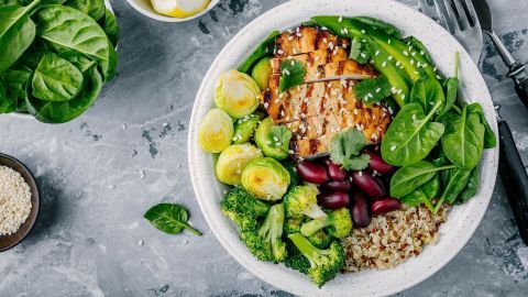 Eine bunte Schüssel mit gesundem Gemüse, Quinoa und Hühnchen, serviert auf einem grauen Untergrund und umgeben von frischem Spinat.