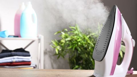 Ein Bügeleisen mit Dampfausstoß steht auf einem Tisch, umgeben von Wäsche und Reinigungsmitteln, während es Dampf abgibt.