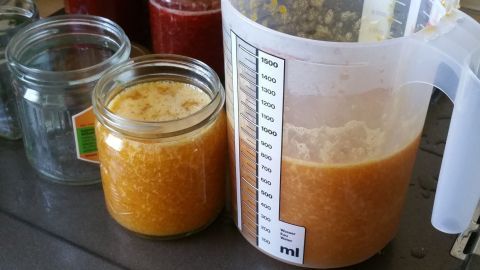 Ein Messbecher mit Marmelade steht neben einem gefüllten Glas auf einer Küchenarbeitsplatte, umgeben von weiteren Gläsern.