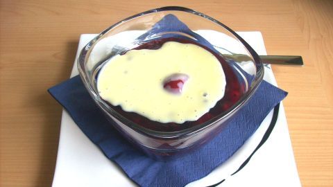 Ein Schälchen Norddeutsche Rote Grütze mit Sahne liegt auf einem weißen Teller und blauen Serviette auf einem Holztisch.