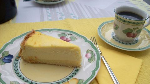 Ein Stück perfekter Käsekuchen liegt auf einem bunten Teller neben einer Tasse Kaffee auf einem gelben Tischset.