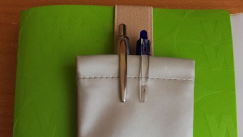 Eine weiße Stiftetasche hängt an einem bunten Notizbuch, bereit, Kulis und Co. für Notizen aufzubewahren.