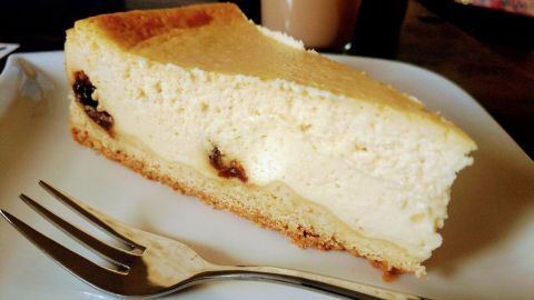 Ein Stück Käsekuchen liegt auf einem weißen Teller, neben einer Gabel, in einem gemütlichen Raum mit einer Tasse Kaffee.
