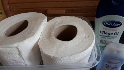 Zwei Rollen Toilettenpapier liegen in einem Behälter, während Pflegeöl und eine andere Flüssigkeit daneben stehen.