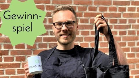Eine Person hält eine Tasse und eine Tasche in der Hand, lächelt und präsentiert die Gewinne eines Gewinnspiels vor einer Backsteinwand.