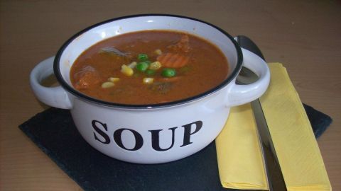 Ein weißer Suppentopf mit der Aufschrift "SOUP" enthält eine kalte, gemüsige Suppe auf einem schwarzen Serviette-Hintergrund.