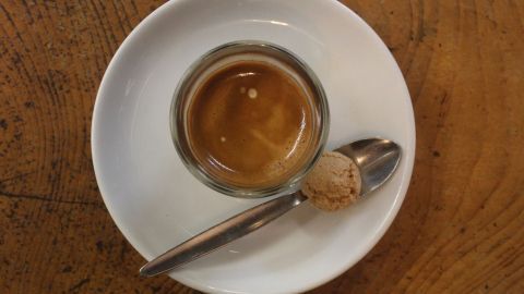 Ein kleiner Espresso in einem Glas steht auf einem weißen Unterteller, daneben liegt ein Löffel auf einem Holztisch.