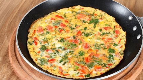 Ein Kräuter-Omelette liegt goldbraun in einer Pfanne, umgeben von einer Holzoberfläche, mit frischen Tomaten und Schafskäse.