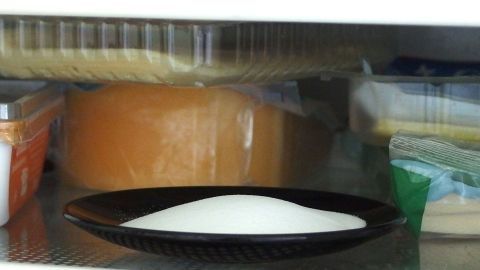 Mit Natron lassen sich wunderbar Gerüche im Kühlschrank oder Geschirrspüler bekämpfen. Ein Teller mit Zucker steht im Kühlschrank, um Gerüche aus anderen Lebensmitteln zu neutralisieren. Der Hintergrund zeigt Käse und andere Lebensmittel.