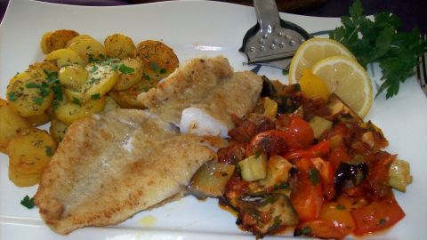 Auf einem weißen Teller liegt zartes Rotbarschfilet neben Ofen-Ratatouille und knusprigen Bratkartoffeln, garniert mit Zitrone.