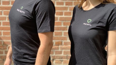 Schwarze T-Shirts mit dem Logo "Frag Mutti" werden von einem Mann und einer Frau an einer Ziegelwand getragen.