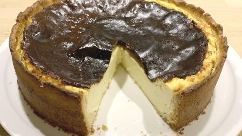Ein Käsekuchen mit Boden und Rand steht auf einem Teller, ein Stück ist herausgeschnitten und auf dem Tisch platziert.