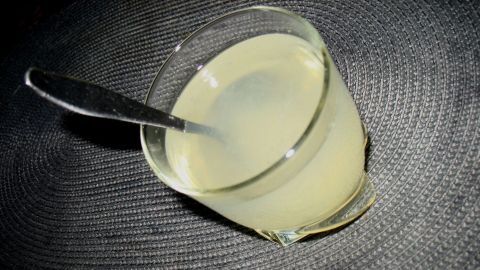 Ein Glas Zitronenwasser steht auf einem dunklen Tisch und reflektiert das Licht, während ein Löffel darin liegt.