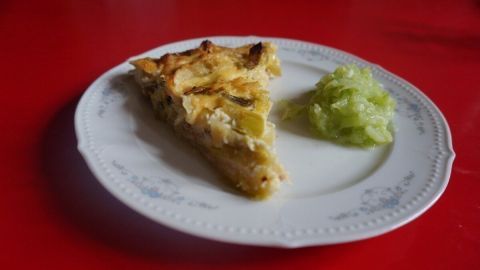 Eine Stück Porree-Torte liegt auf einem weißen Teller neben einem Klecks grüner Beilage, alles auf rotem Tisch.