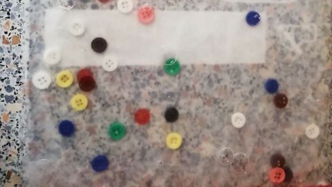Bunte Knöpfe liegen in einem transparenten Beutel, während kleine Kinder sie mit den Händen erkunden.