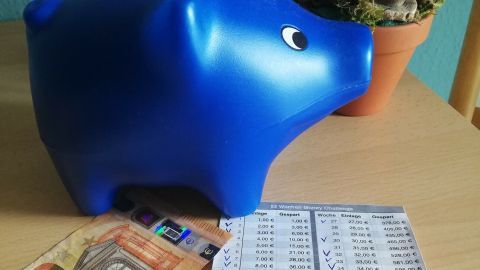 Ein blauer Sparschwein steht auf einem Tisch, neben ihm liegt ein 50-Euro-Schein und eine Liste mit Sparbeträgen.