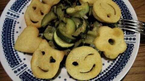 Bunte Emoti-Potatoes lächeln auf einem Teller, umgeben von einer frischen Gemüsepfanne aus Zucchini und anderen Gemüsesorten.