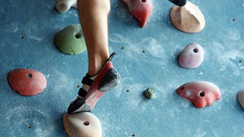 Ein Fuß in speziellen Kletterschuhen tritt auf einen bunten Griff an einer Innenkletterwand in einem Sportzentrum.