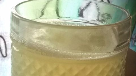 Ein transparentes Glas enthält eine gelbliche Flüssigkeit, in der Honig zu lösen scheint, vor einem gemusterten Hintergrund.