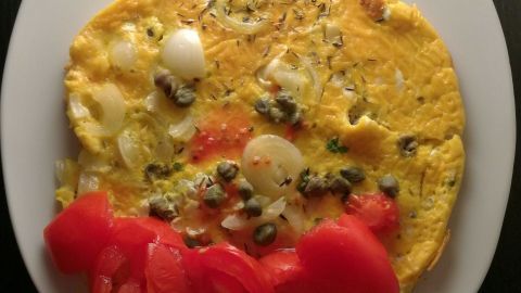 Eine bunte Frittata liegt auf einem weißen Teller, garniert mit frischen Tomatenscheiben und Kräutern.