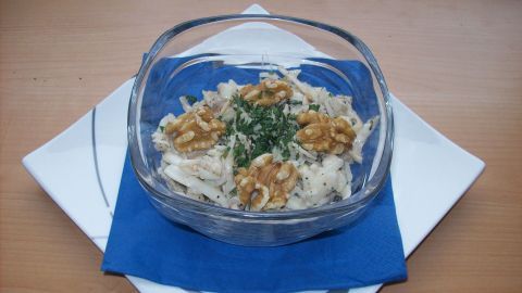 Ein klarer Glasschüssel enthält Waldorf-Salat mit Äpfeln und Nüssen, auf einem weißen Teller mit blauer Serviette.