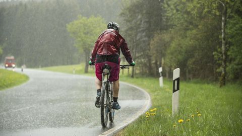 Ein Radfahrer fährt bei Regen auf einer Landstraße vorbei an grünen Wiesen und Bäumen, was sportliche Aktivitäten bei schlechtem Wetter zeigt.