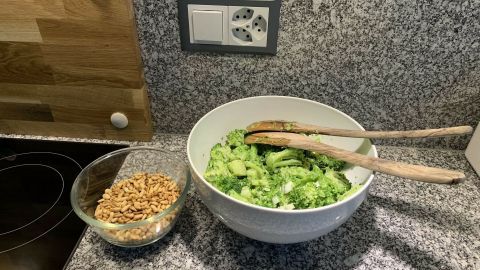 Ein frischer Brokkolisalat liegt in einer großen weißen Schüssel auf einer grauen Arbeitsplatte, daneben eine Schale mit gerösteten Nüssen.