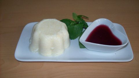Ein Grießpudding liegt auf einem weißen Teller neben einer herzförmigen Schale mit Himbeersirup und frischen Minzblättern.