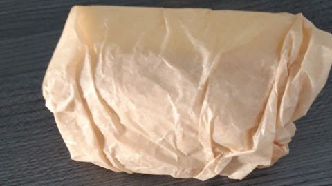 Ein Stück Käse ist in dünnem Papier eingewickelt und liegt auf einem dunklen Holzoberfläche, bereit zur Aufbewahrung.