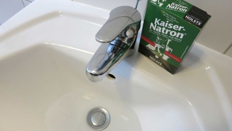 Ein Vorschlag bei Empfindlichkeit gegen Seife: Gesicht mit einer Speisenatron-Wasserlösung gründlich waschen, ohne mit klarem Wasser nachzuspülen. Ein glänzender Wasserhahn befindet sich über einem Waschbecken, während eine Packung mit Kaisernatron daneben liegt.