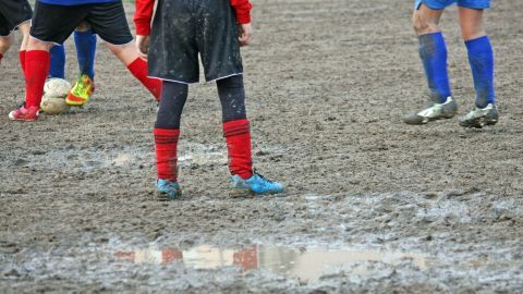 Auf einem matschigen Fußballfeld stehen Kinder in roten und blauen Trikots, während sie im Regen spielen.