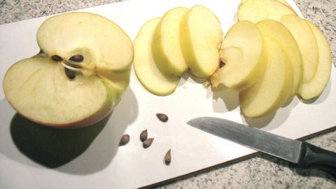 Auf einem Schneidebrett liegt ein halbierter Apfel, umgeben von geschnittenen Apfelstücken und Apfelkernen. Eine Klinge schneidet den Apfel.