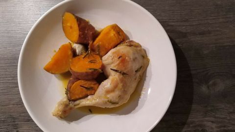 Saftige Hähnchenkeulen liegen zusammen mit gebackenen Süßkartoffeln auf einem weißen Teller auf einem dunklen Tisch.