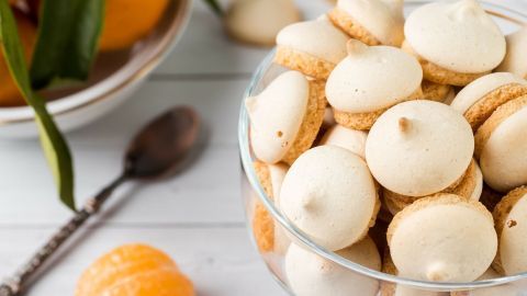 Zarte Baiser Plätzchen liegen in einer Glas-Schüssel, umgeben von frischen Orangen und einem Löffel auf einem weißen Holztisch.