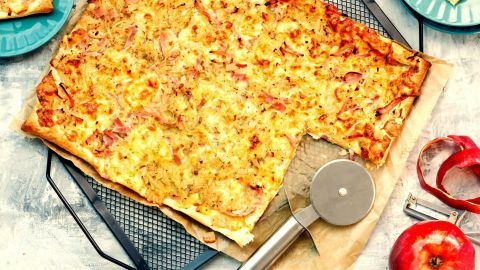 Eine frisch gebackene Blätterteigpizza liegt auf einem Kuchengitter und wird mit einem Pizzaschneider geschnitten.