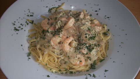 Riesengarnelen und Lachswürfel liegen in einer cremigen Kräutersahnesauce auf einem Bett aus Spaghetti, mit frischen Kräutern bestreut.