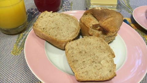 Ein DDR-Brötchen liegt auf einem rosa Teller, umgeben von Butter, Marmelade und einem Glas Saft, bereit zum Frühstück.