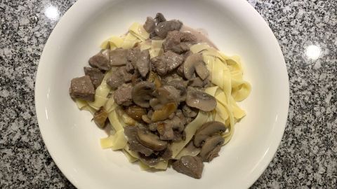 Eine Portion Rindsragout liegt auf Tagliatelle in einer weißen Schüssel, angerichtet auf einer grauen Marmoroberfläche.