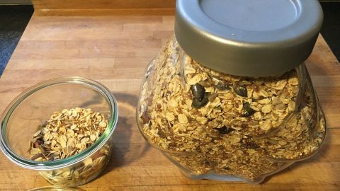 Eine große, offene Gläser mit selbst gemachtem Müsli steht auf einem Holzbrett, daneben ein kleinerer Behälter mit Müsli.