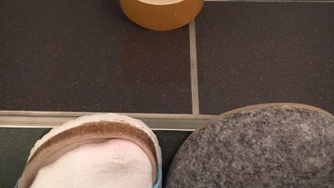 Hausschuhe liegen neben einem Klebestreifen auf einem gefliesten Boden, bereit zum Kleben der gelösten Sohlen.