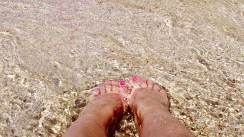 Mithilfe von Deokristallen kann man auch Schweißfüße hervorragend bekämpfen. Füße stehen im klaren Wasser, das sanft um sie plätschert. Der Kontext ist ein entspannender Strandtag, ideal gegen Schweißfüße.