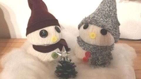 Zwei Schneemannpärchen aus alten Socken stehen auf einer Platte in einer weichen, weißen Umgebung und umgeben von Deko.