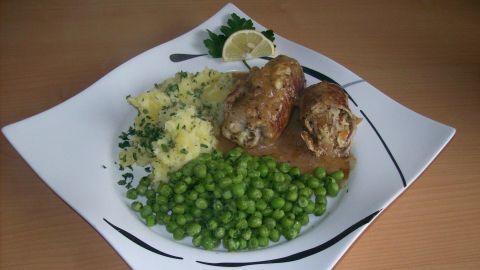 Saftige Schweinsrouladen auf einem Teller, begleitet von grünen Erbsen und Kartoffelsalat, alles appetitlich angerichtet.