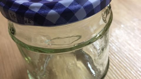 Ein Glas mit einem karierte Deckel steht auf einem Holztisch, bereit zur Etikettenentfernung für die Wiederverwendung.