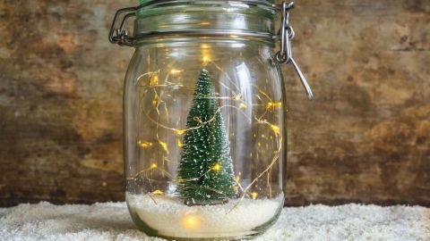 Ein Weckglas enthält einen kleinen Tannenbaum, umgeben von Lichtern auf weißem Schnee, in festlicher, winterlicher Atmosphäre.