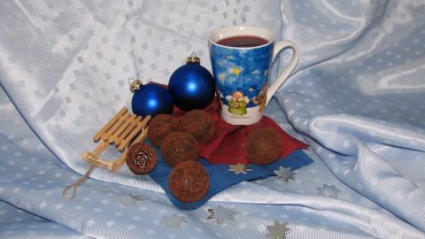 Eine festlich dekorierte Tasse steht auf einem blauen Tuch, umgeben von blauen Kugeln, Äpfeln und einem Holzschlitten.