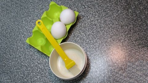 Zwei frische Eier liegen in einem grünen Eierkarton, während eine gelbe Beschichtungsspritze in einer weißen Schüssel mit Eiweiß steht.