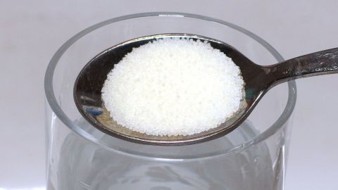 Ein Löffel mit weißem Zuckerkristallen schwebt über einem Glas Wasser und könnte als Hausmittel gegen entzündetes Zahnfleisch dienen.
