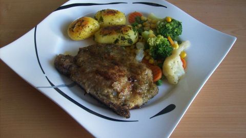Ein würziges Kotelett mit Kräuter-Panade liegt auf einem Teller, umgeben von Gemüse und Bratkartoffeln, appetitlich angerichtet.